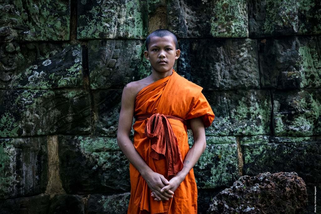 Monk in bright orange robe stands in front of the green ruins of Angkor Wat