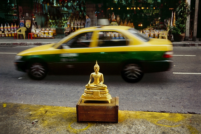 A mini buddha statue spotted with a local Thailand taxi as background - Photo by Steve McCurry